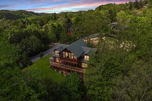 Eine große, mehrstöckige Blockhütte mit umlaufender Veranda, umgeben von dichtem Wald und Hügeln.