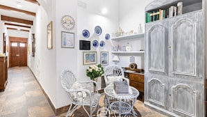 Een hal met witte muren, een houten deur en een witte rieten stoel. Er hangen blauwe en witte borden aan de muur, een witte vaas met bloemen en een grijze kast met boeken.