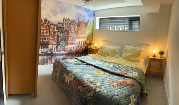 Un dormitorio con cama, mesita de noche, lámpara de pared y ventana con persianas.