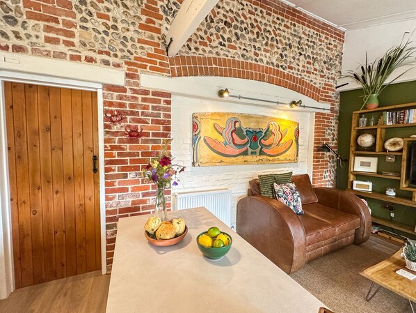 A cozy living room with a brick wall, a wooden door, a brown leather sofa, a table with a bowl of biscuits and lemons, and a green bookshelf.