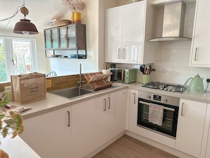 A modern kitchen with white cabinets, a gas stove, a built-in oven, a microwave, and a toaster. There is a window providing natural light and a hanging light fixture. - Nicholson Cottage  - sleeps 4 guests  in 2 bedrooms (Happisburgh)
