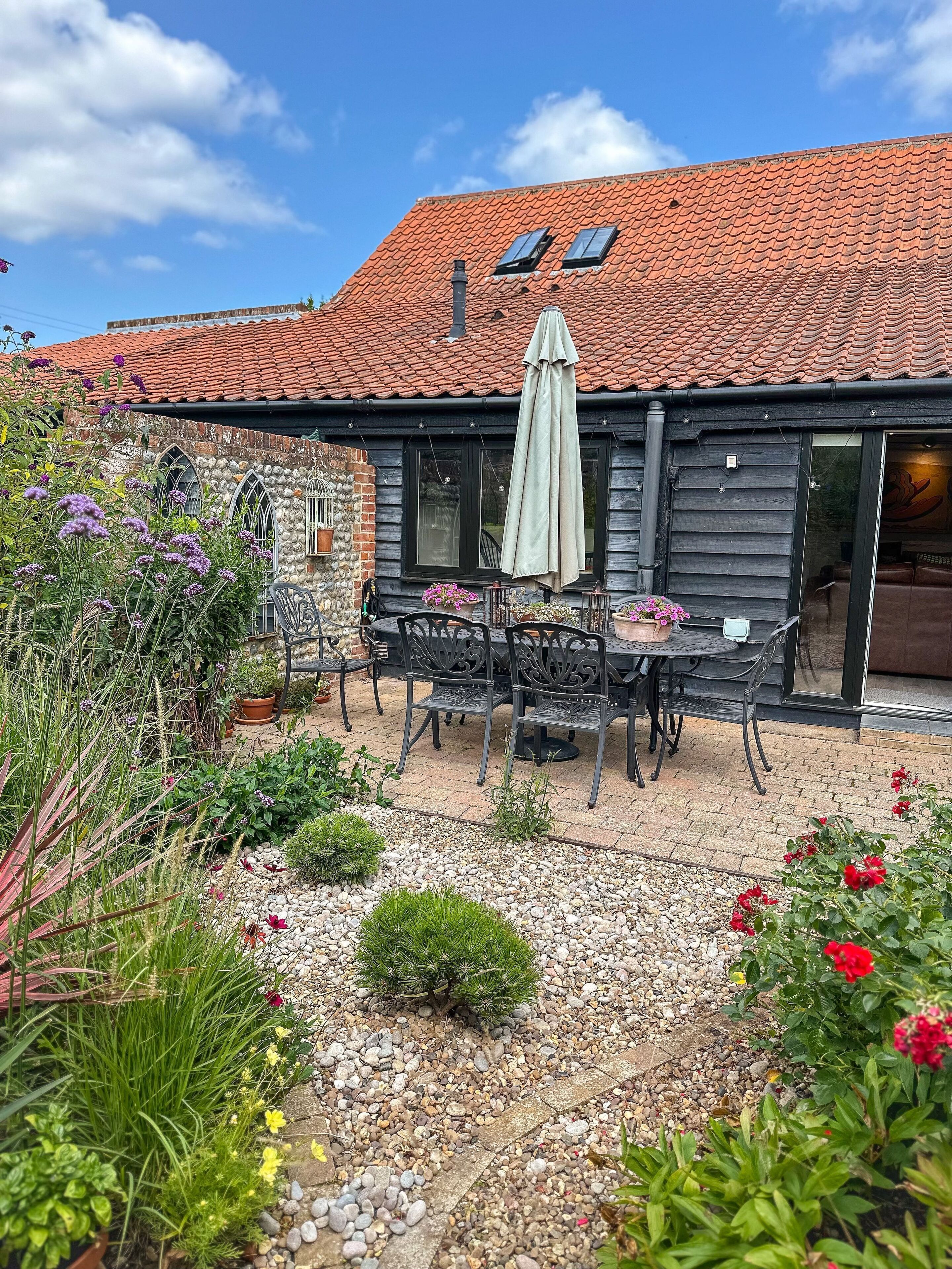 Un bâtiment rustique avec un toit en tuiles, une terrasse avec une table et des chaises, et un jardin agrémenté de diverses plantes et fleurs.