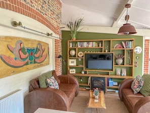 A cozy living room with a fireplace, leather sofas, a wooden coffee table, and a bookshelf filled with books and decorative items. - Nicholson Cottage  - sleeps 4 guests  in 2 bedrooms (Happisburgh)