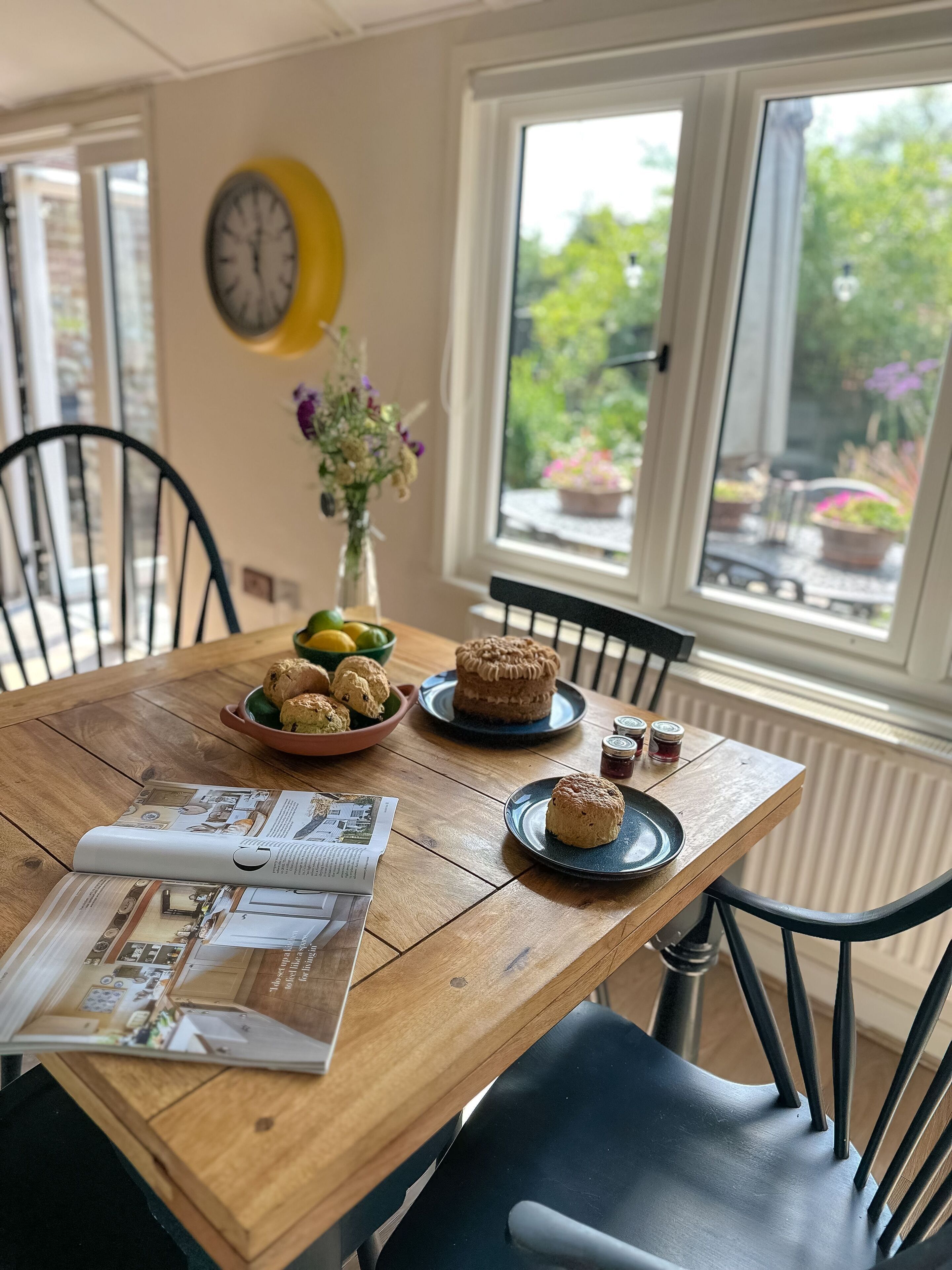 A wooden dining table with a magazine, a bowl of cookies, a cake, and a vase of flowers.