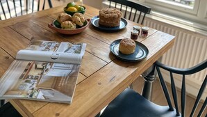 Un tavolo da pranzo in legno con una rivista, una ciotola di biscotti, una torta e un vaso di fiori.