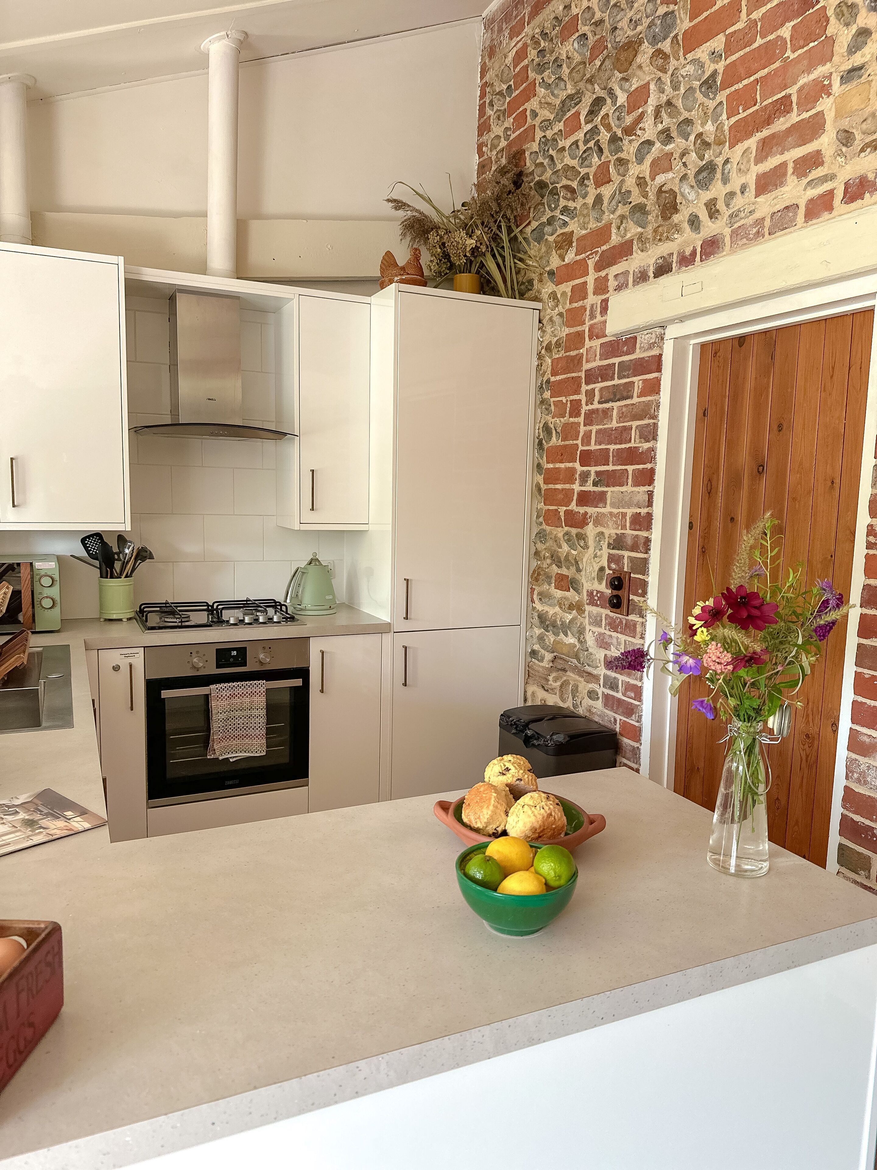 Une cuisine avec des armoires blanches, un mur en briques, une porte en bois et un vase rempli de fleurs posé sur le comptoir.