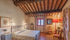 Een slaapkamer met een bed, een kaptafel, een stoel, een tafel met een lamp en een raam met gordijnen.