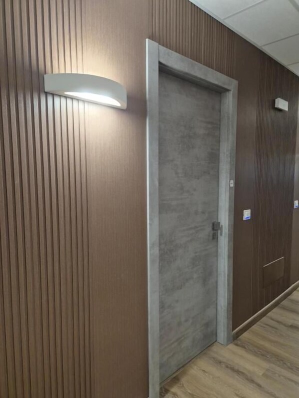 Un couloir aux murs recouverts de lambris en bois, une porte grise et un luminaire placé au-dessus.