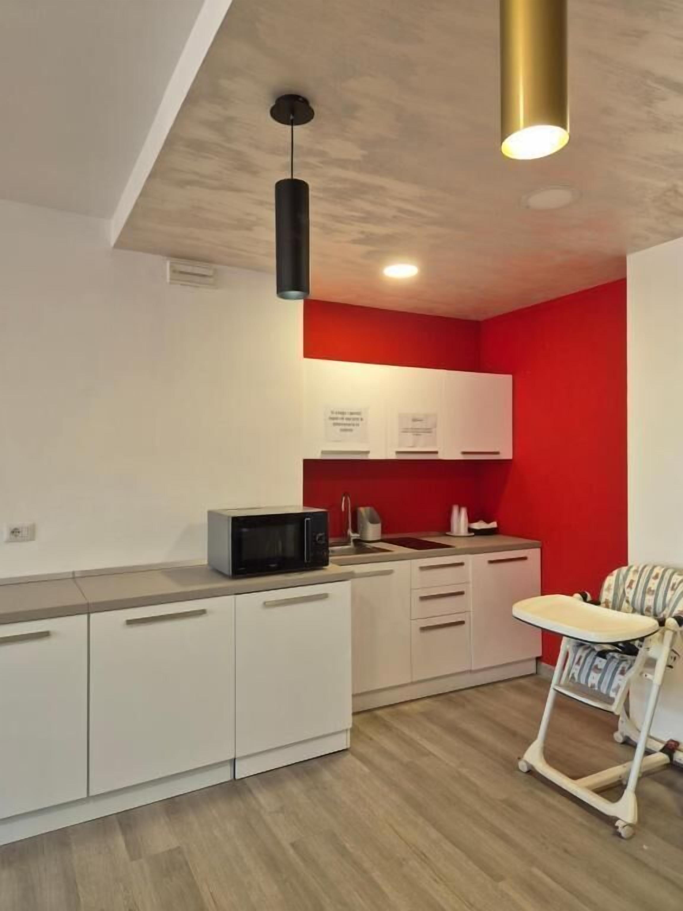 A modern kitchen with white cabinets, a red accent wall, and a small dining area with a high chair.