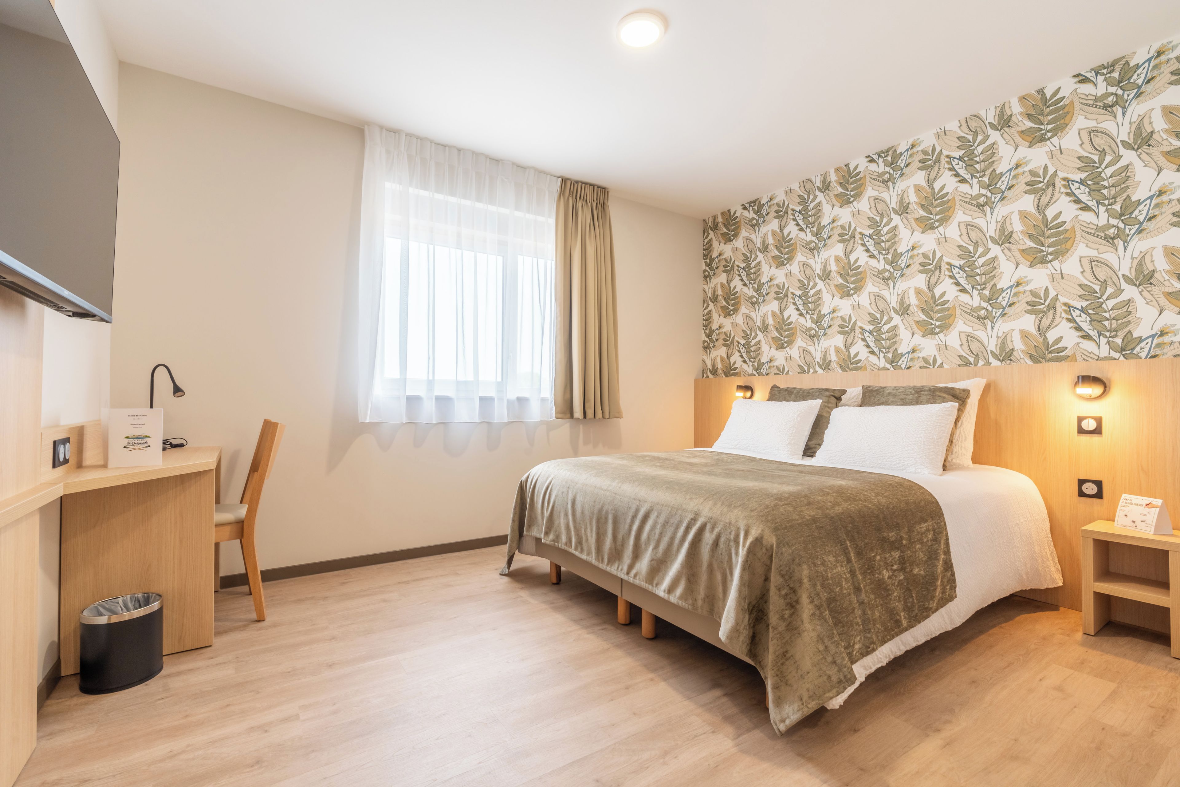 Une chambre d’hôtel comprenant un lit, un bureau, une chaise, une poubelle, une fenêtre avec des rideaux et un mur orné d’un motif de feuilles.