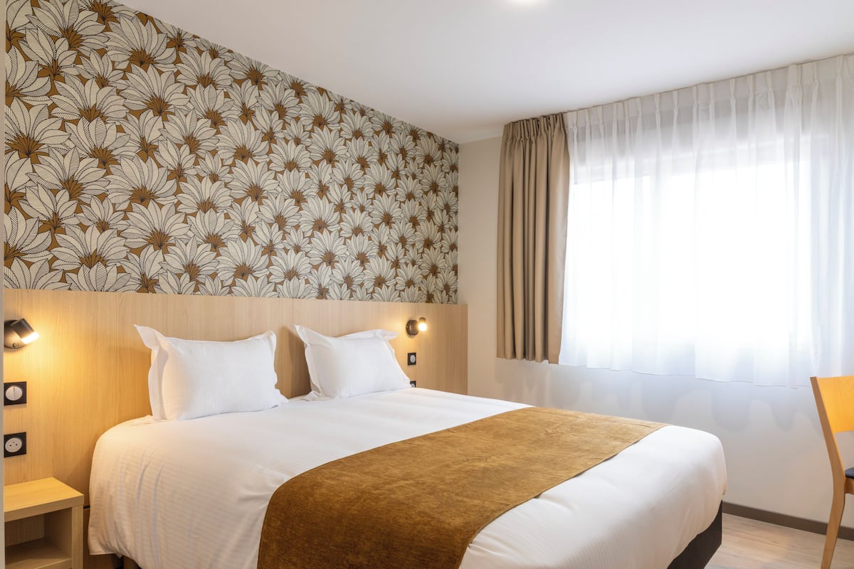 A hotel room with a large bed, a headboard with a floral pattern, a bedside table, and a window with curtains.