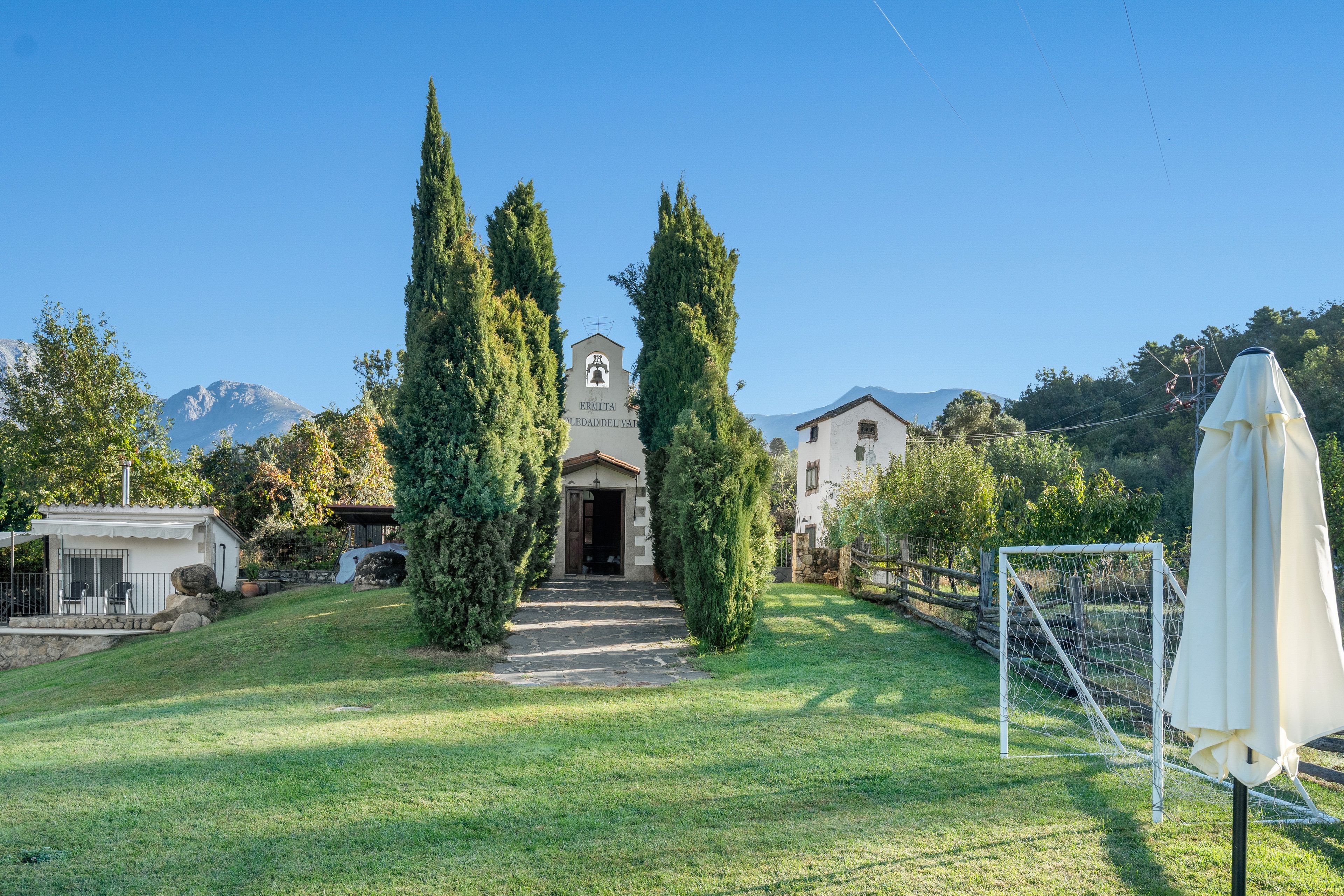 A residential property with a stone pathway leading to a white building with a bell tower, surrounded by tall cypress trees and a mountain backdrop.