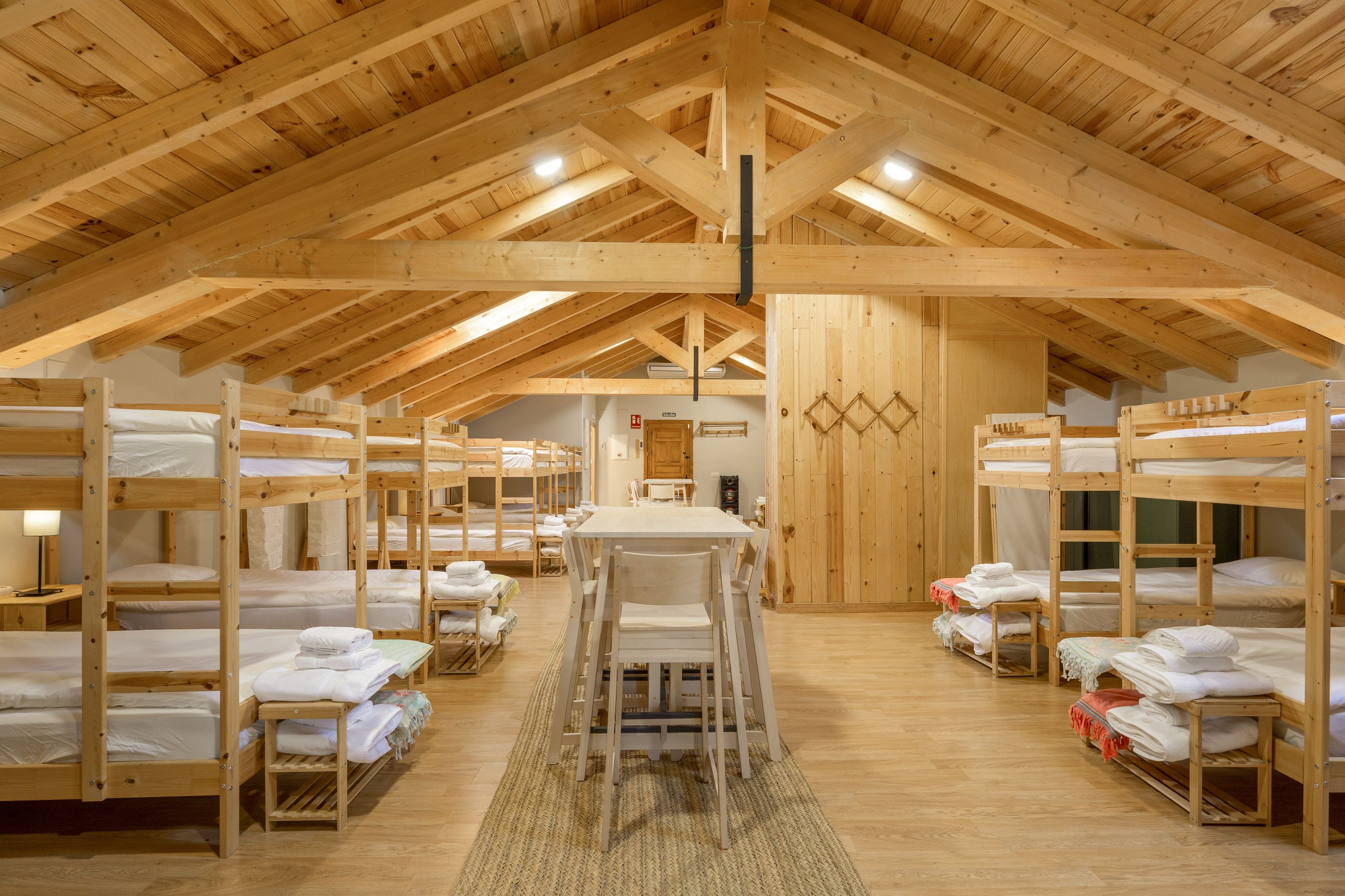 Un dormitorio con literas dispuestas en filas, una mesa central y un alto techo de madera con vigas a la vista.