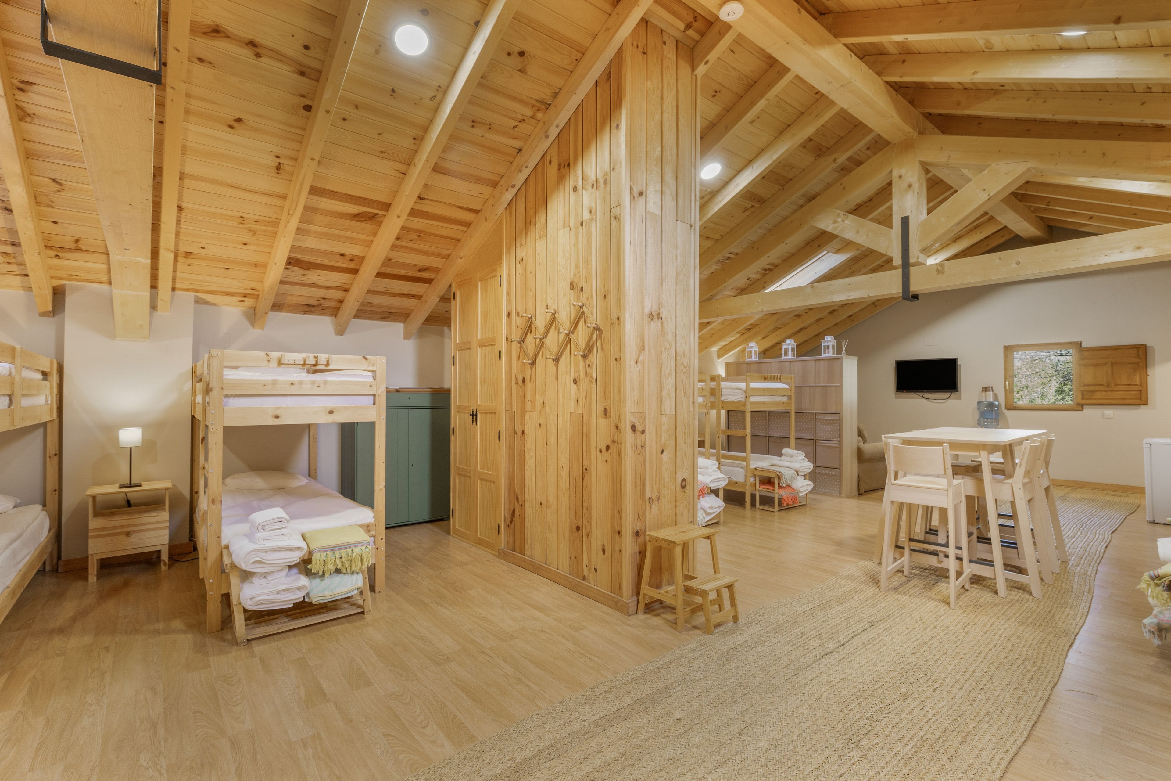Una habitación con techos altos, vigas de madera, literas, una mesa con sillas y un televisor.