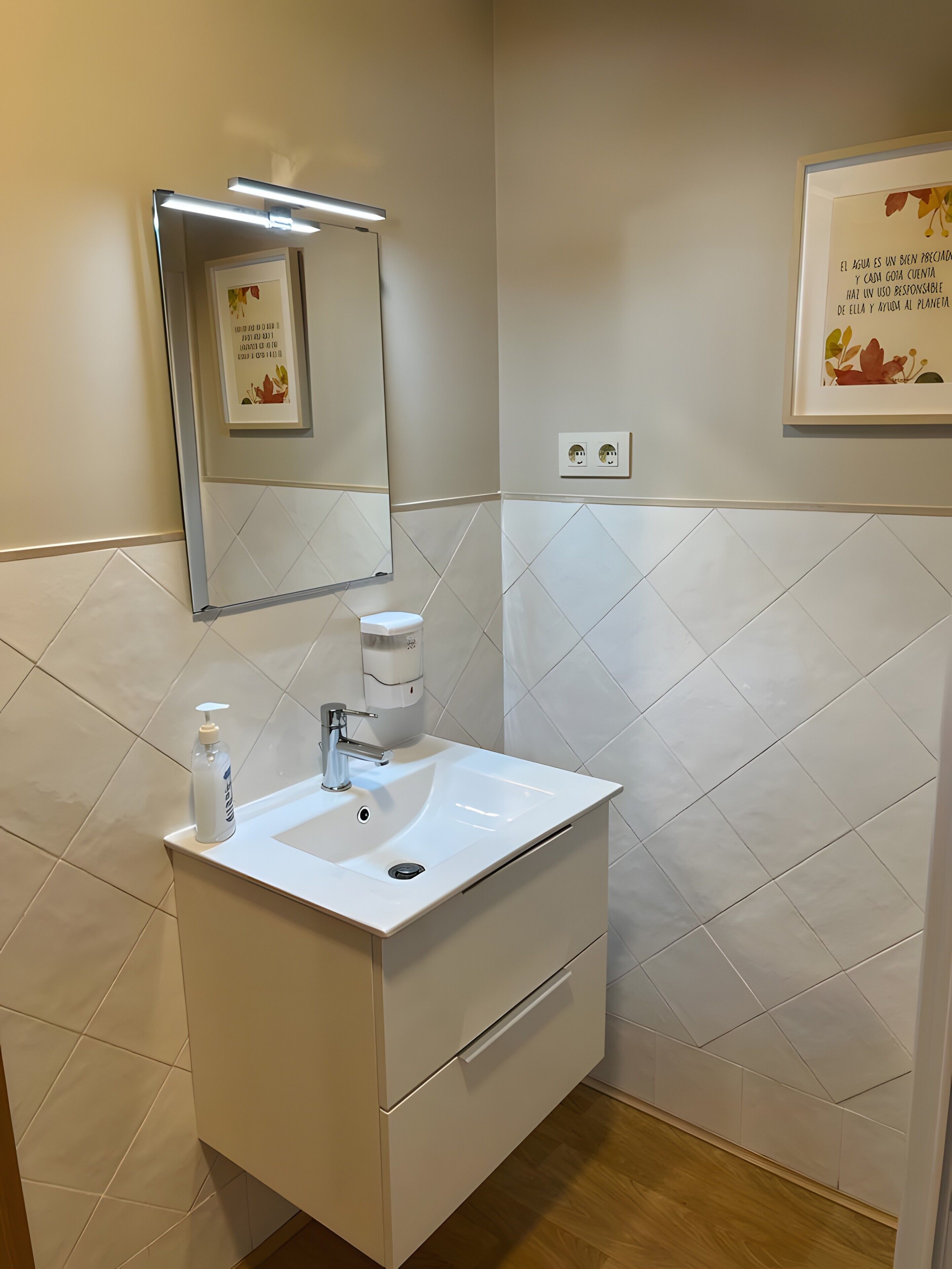 Een badkamer met een wit wastafel, een spiegel, een zeepdispenser en een lijst met een citaat aan de muur.