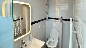 A modern bathroom with a glass shower enclosure, a white toilet, and a vanity with a mirror. - Royal Crown Apartment With Royal Garden (Le Bourget)