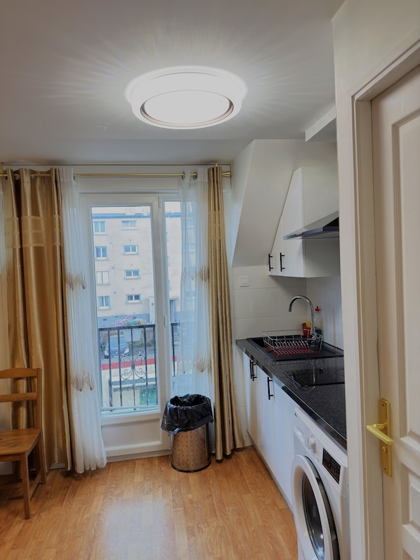 A compact kitchen with a washing machine, a sink, and a window with curtains. - Royal Crown Apartment With Royal Garden (Le Bourget)