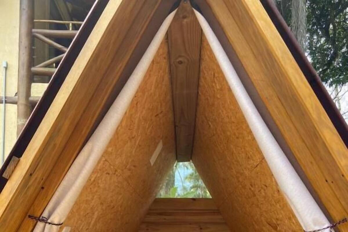 A small, triangular wooden tent with a bed, a fan, and a view of trees through a window.