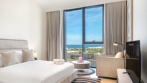 A hotel room with a large bed, a desk, a chair, a TV, and a view of the sea.