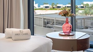 A hotel room with a bed, a round table with a vase of flowers, and a view of the ocean and buildings.