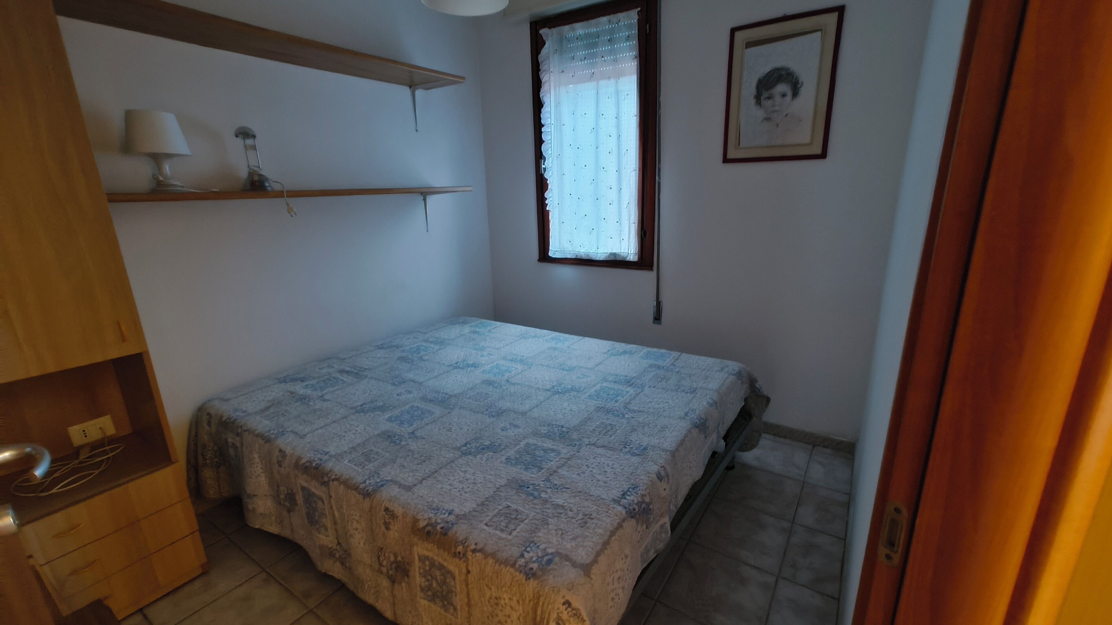 A small, clean room with a single bed, a window with a curtain, a shelf with a lamp, and a framed picture on the wall.