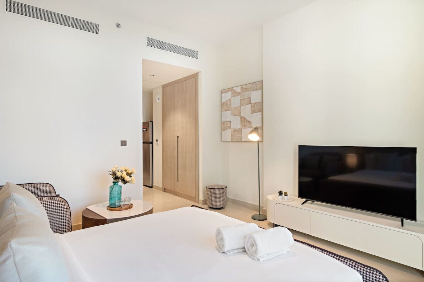 Een moderne hotelkamer met een groot bed, een flatscreen televisie, een klein nachtkastje met een vaas, en een gang die naar een deur leidt.