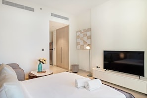 A modern hotel room with a large bed, a flat-screen TV, a small bedside table with a vase, and a hallway leading to a door. - FIRST CLASS | 3BR | Stunning Beachfront Bliss (Al Riffa)