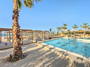 A poolside area with lounge chairs, a palm tree, and a clear blue sky. - FIRST CLASS | 3BR | Stunning Beachfront Bliss (Al Riffa)