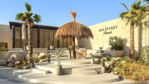 A modern resort with a central thatched structure, palm trees, and a stone wall with the name 'BAR DU PORT Beach'. - FIRST CLASS | 3BR | Stunning Beachfront Bliss (Al Riffa)
