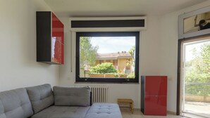A living room with a grey sectional sofa, a small wooden stool, a red cabinet, and a window with a view of a house and greenery. - Atelier Vella by Interhome (Scarlino)