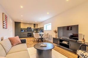 A modern living room with a sofa, a coffee table, and a TV. There is a kitchen in the background with a dining area. - Stylish Flat, Family Friendly, Walk to The Oracle (Reading)