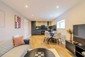 Un salon moderne avec un canapé, une table basse et un meuble pour la télévision. On y trouve également un coin repas avec une table et des chaises. La cuisine, équipée d’appareils électroménagers et de meubles modernes, est visible au fond.