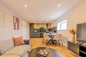 A modern living room with a sofa, a coffee table, and a dining area with chairs. A kitchen is visible in the background with a TV on a stand. - Stylish Flat, Family Friendly, Walk to The Oracle (Reading)
