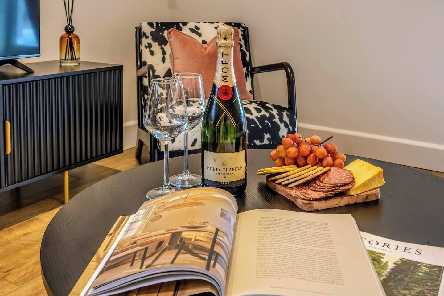 Une table avec une bouteille de champagne Moët & Chandon Brut, deux verres à vin, un livre et une assiette de fromage et de raisins.