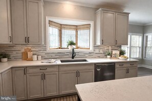 A kitchen with light gray cabinets, a white countertop, a built-in dishwasher, and a window with blinds. - Spacious 6bed Woodland Retreat With Movie Room Deck (California)