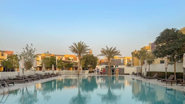 A swimming pool with lounge chairs and umbrellas, surrounded by buildings and trees.