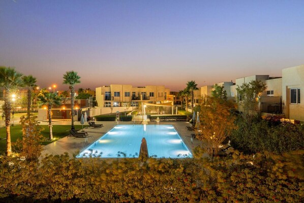 A swimming pool with lounge chairs and palm trees, surrounded by modern buildings at dusk.