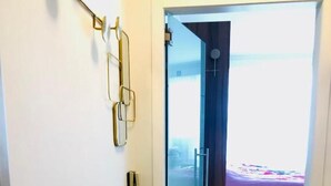 A narrow hallway with a fire extinguisher on the left, a glass door leading to a bedroom with a bed and a window. - Studio Oostende Dichtbij Strand Centrum (Ostend)
