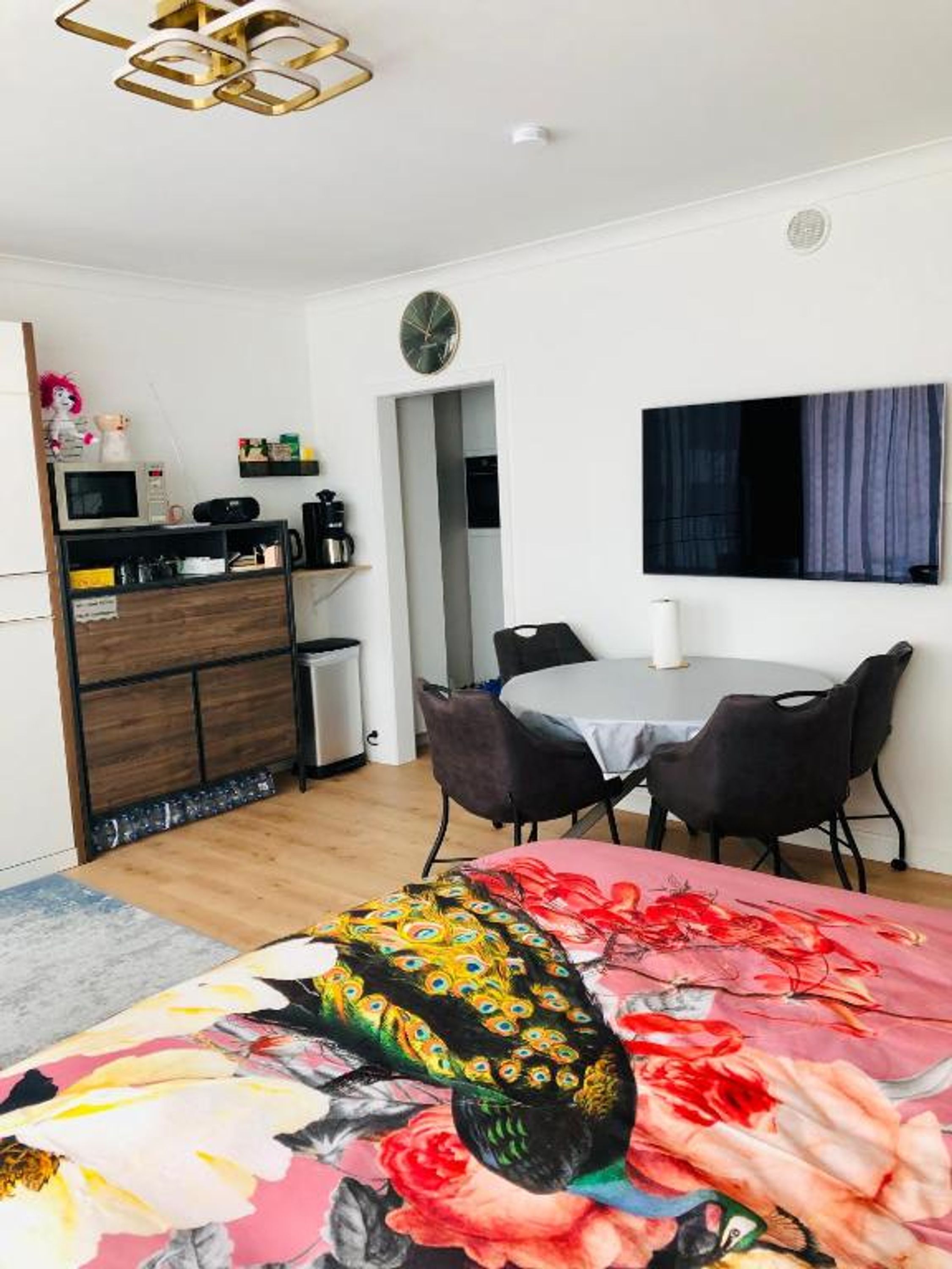 A modern room with a bed, a dining table with chairs, a TV mounted on the wall, and a kitchenette area.