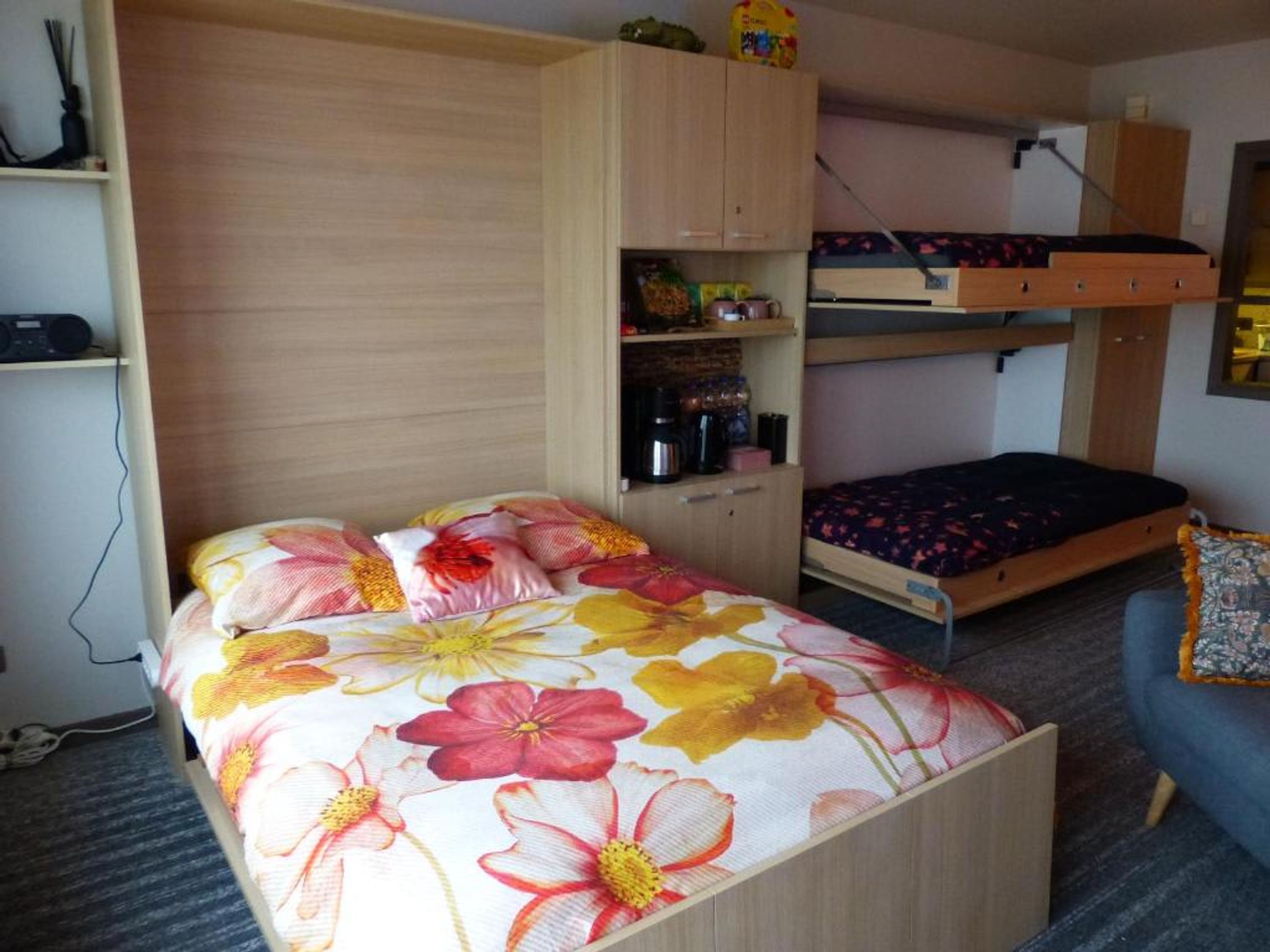 Une petite pièce comprenant un lit superposé, un lit simple avec une literie à motifs floraux, une petite kitchenette et une chaise.