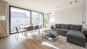 A modern living room with a grey sofa, a coffee table, and a dining area with chairs and a table. - Levant Appartement 2 Slaapkamers 2 Badkamers Balkon Knokke-heist (Knokke-Heist)