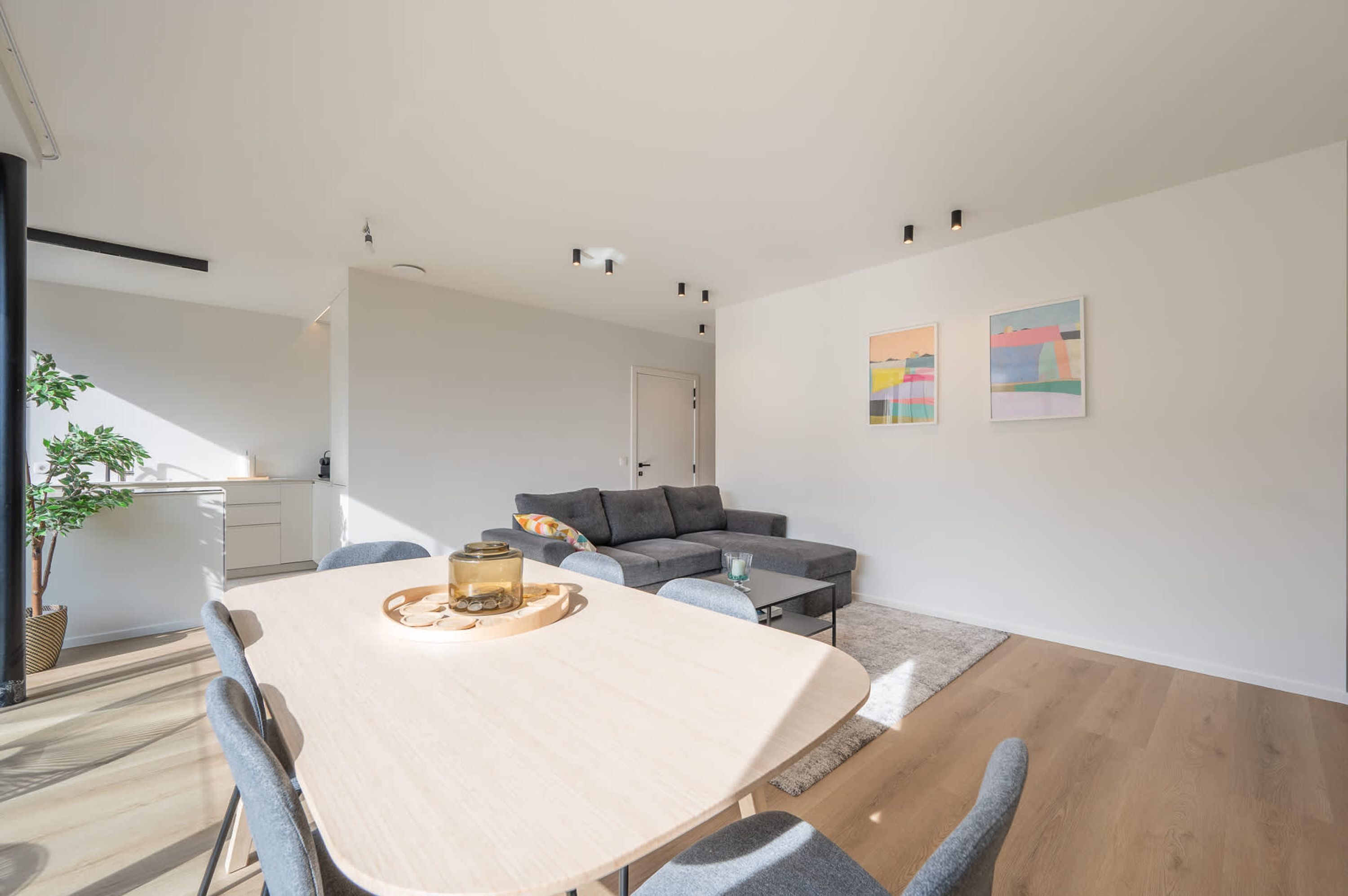 A modern living room with a grey sofa, a dining table with chairs, and a staircase leading to an upper floor.