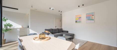 A modern living room with a grey sofa, a dining table with chairs, and a staircase leading to an upper floor.