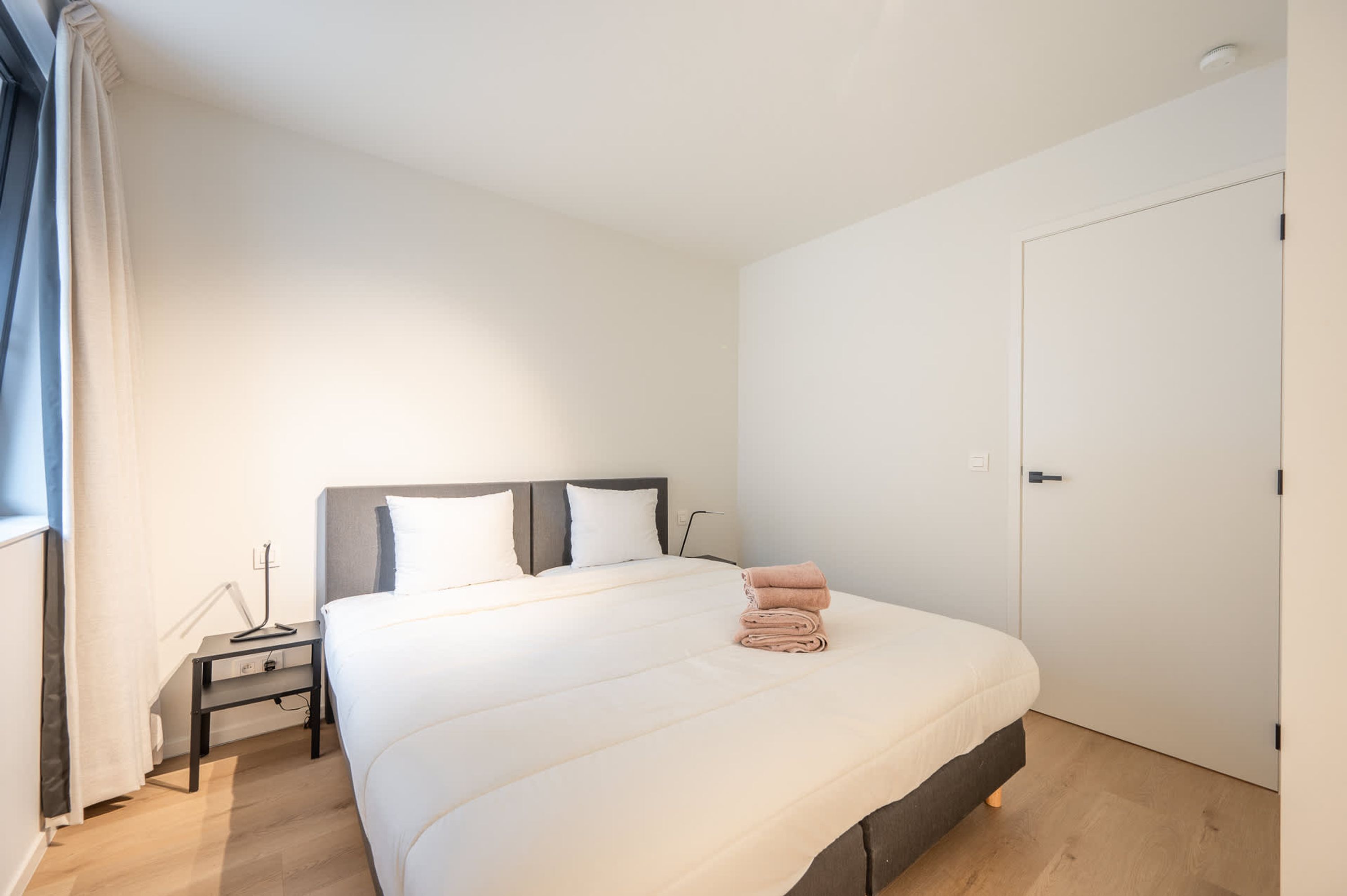 A neatly made bed with a folded towel, a bedside table with a lamp, and a door to the right.