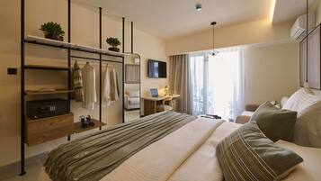 A hotel room with a bed, a desk with a laptop, a TV, a plant, and a wardrobe.