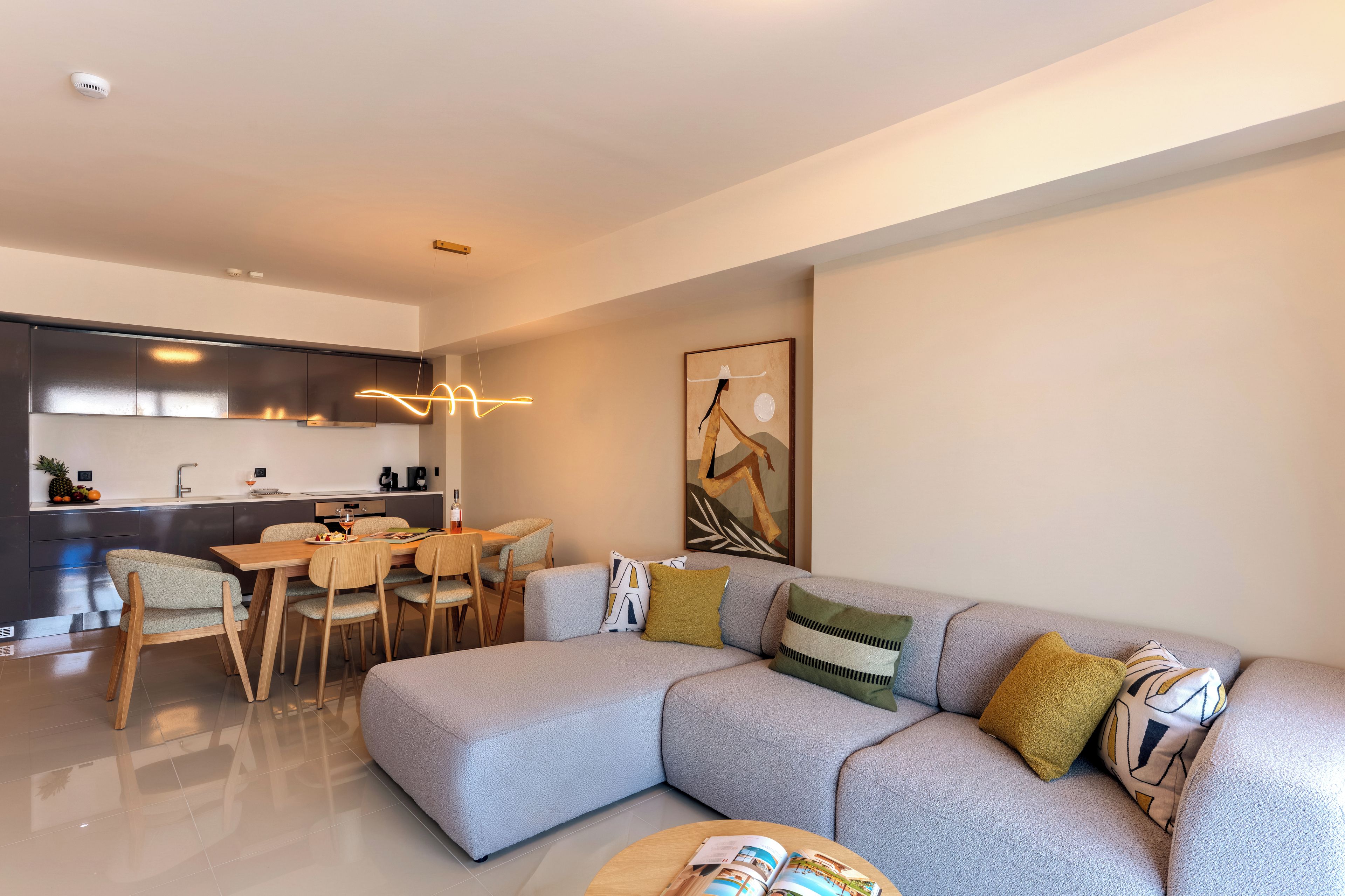 Un salon moderne avec un canapé d’angle, un coin repas avec des chaises, une cuisine dotée d’une hotte en acier inoxydable et un luminaire suspendu.