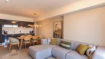 Un salon moderne avec un canapé d’angle, un coin repas avec des chaises, une cuisine dotée d’une hotte en acier inoxydable et un luminaire suspendu.