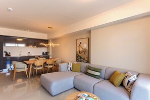 Una sala de estar moderna con un sofá modular, un comedor con sillas, una cocina con extractor de acero inoxidable y una lámpara colgante.