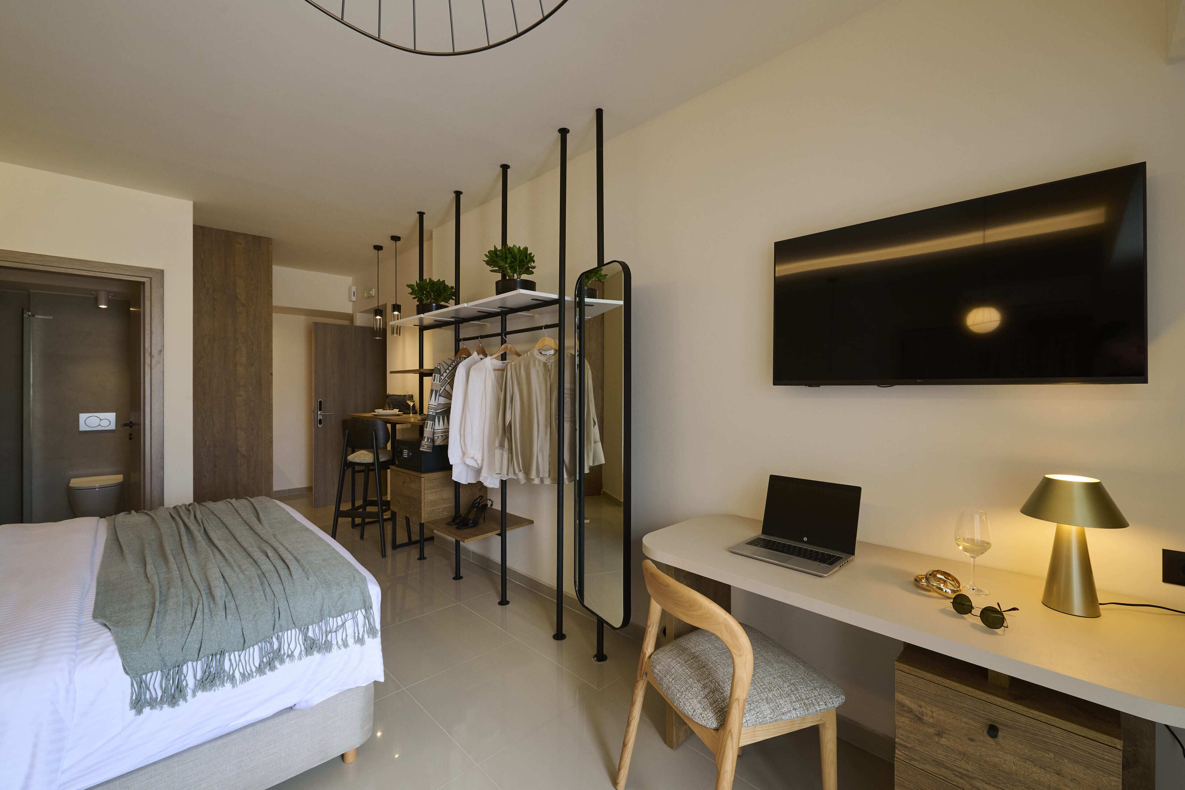 A hotel room with a bed, desk, laptop, chair, TV, and a small table with a plant and clothes.