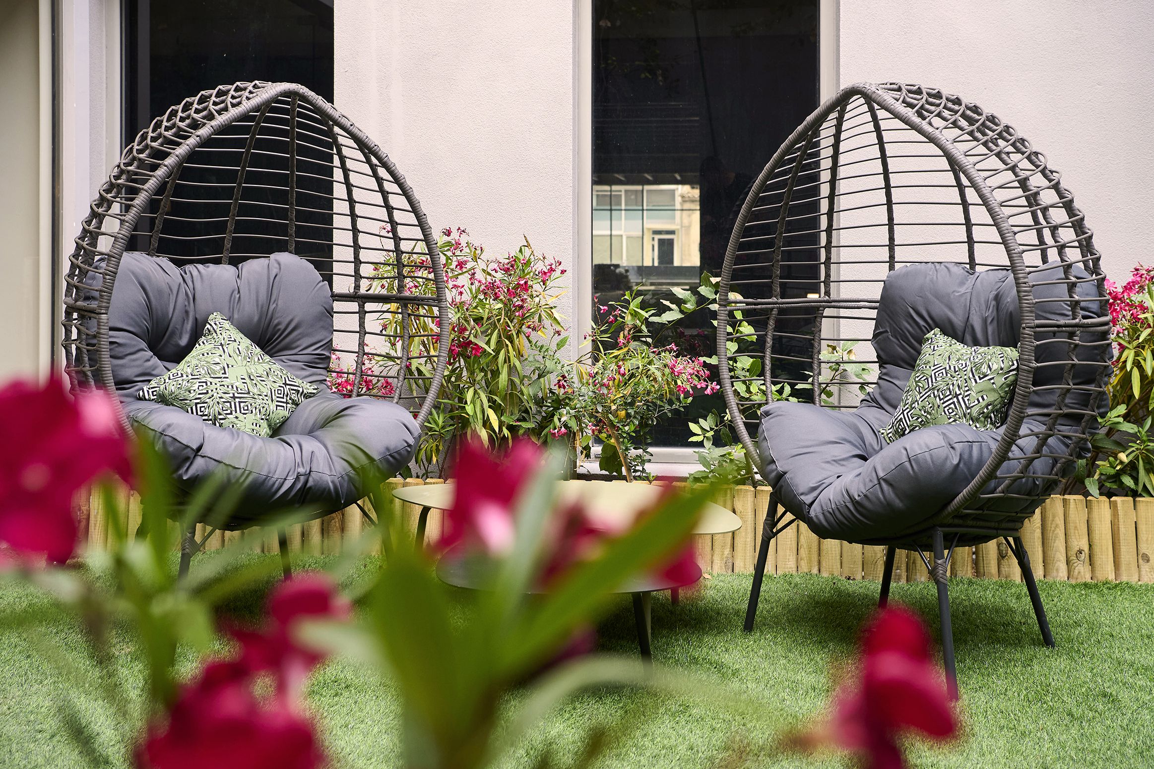 Deux fauteuils en osier avec des coussins, une petite table et un pot de fleurs orné de fleurs roses.
